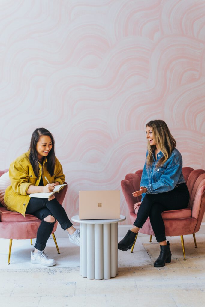 two women sitting together in suede chairs, one is writing, both are laughing